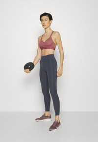 Reggiseno sportivo borgogna, leggings blu navy a vita alta e scarpe da ginnastica scure. Modella che tiene un disco di peso nero. Tessuti lisci e attillati.