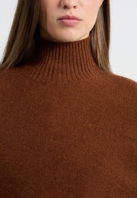 Brązowy sweter z golfem wykonany z miękkiego materiału o prążkowanej fakturze. Ubranie charakteryzuje się wysokim kołnierzem i prostym designem bez dodatkowych wzorów.