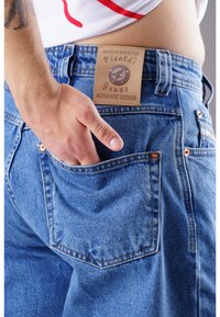 Hellblaue Jeans aus Denim mit einer abgenutzten Textur, versehen mit einem Lederschild, das "Picaldi Jeans Authentic Design" beschriftet ist, und einer Hand in der Gesäßtasche.