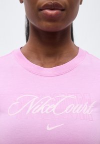 Κοντινό πλάνο ενός ατόμου που φοράει ροζ πουκάμισο με λευκό κείμενο «Nike Court» και μικρό λογότυπο Nike swoosh κάτω από το κείμενο.