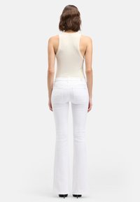 Jeans bianchi a zampa con una texture liscia e tasche posteriori; abbinati a un top beige chiaro senza maniche. Il look è completato da décolleté nere.