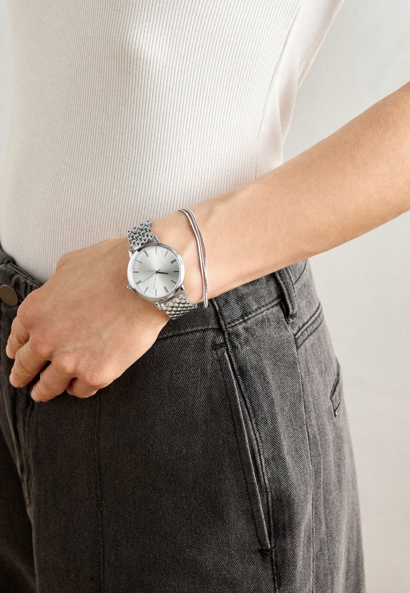 Silverfärgad rund klocka med nätarmband, som visar en minimalistisk urtavla. Buren på en hand med ett silverarmband, mot mörk tyg.