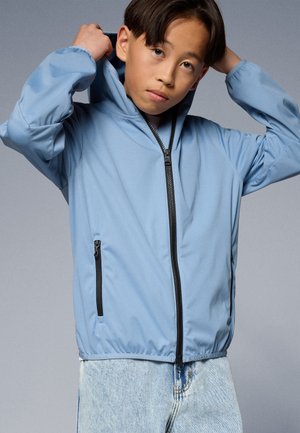 Veste zippée bleu clair avec capuche, dotée de deux poches zippées noires et de poignets élastiques, portée par-dessus un jean en denim clair.