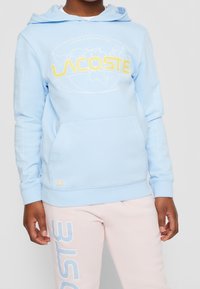 Ljusblå hoodie med en jordglobsgrafik och gul "LACOSTE"-text. Har en känguruficka och rutmönster på ärmarna. Mjuk tyg.