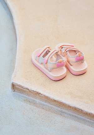 Sandali per bambini rosa e grigi con doppie cinghie in velcro, suole testurizzate e un logo giocoso, posati su una superficie sabbiosa chiara.