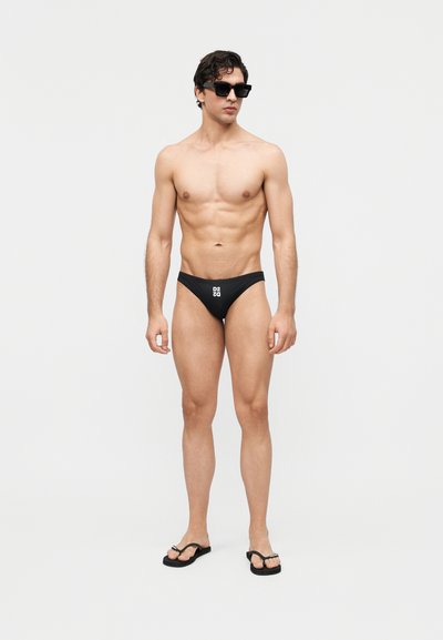 Άντρας χωρίς πουκάμισο, φοράει μαύρο μαγιό με λευκό λογότυπο, μαύρα γυαλιά ηλίου και μαύρα σαγιονάρες, στέκεται μπροστά σε απλό λευκό φόντο.