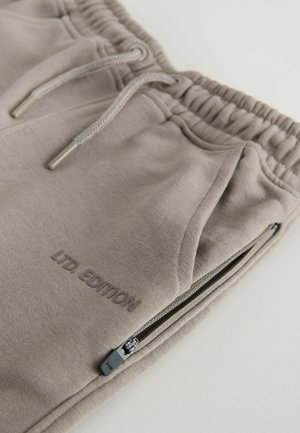 Gaiši bēšu sporta bikses, kas izgatavotas no mīkstas auduma, ar elastīgu jostu, auklām un kabatām ar rāvējslēdzējiem. Redzams teksts "LTD EDITION".