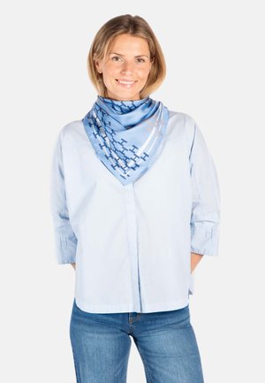 Smilende kvinne iført lyseblå skjorte med brodert H på ermene og blå mønstret silkeskjerf, kombinert med blå jeans.