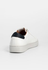 Basket basse blanche avec languette de talon noire, semelle blanche et intérieur marron clair, vue en trois-quarts arrière sur fond blanc.
