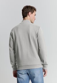 Man die met zijn rug naar de camera staat, gekleed in een lichtgrijze zip-up jack en blauwe denim jeans, tegen een effen lichte achtergrond.