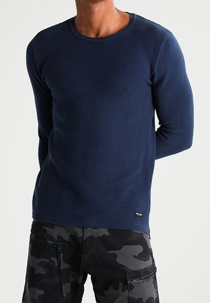 Mann trägt einen marineblauen, strukturierten Pullover mit langen Ärmeln und schwarz-graue Camouflage-Cargohosen, steht mit den Händen hinter dem Rücken.