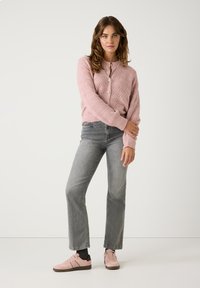 Hellrosa gestrickte Strickjacke mit einem strukturierten Muster und Knopfverschluss; kombiniert mit grauen gerade geschnittenen Jeans und hellrosa Turnschuhen.