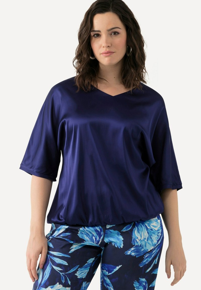 Blusa in satin blu navy con scollatura a V, maniche corte e vestibilità ampia, abbinata a pantaloni blu a fantasia floreale con sfondo scuro.