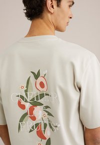 Beige pamut póló kerek nyakkal. A hátsó részén színes virágmotívum és a "CUVILLER LES FRUITS" felirat található. Rövid, laza ujjakkal.