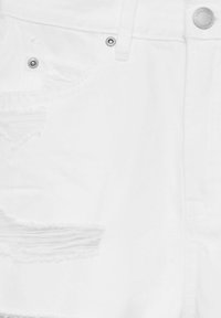 Shorts en denim blanc avec des zones déchirées, des éléments en métal argenté et une fermeture à bouton. Le tissu a une texture lisse avec des bords effilochés sur les poches.