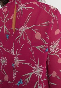 Blus i genomskinligt magentafärgat tyg med blommönster i gröna blad samt rosa och blå blommor. Nyckelhålsringning bak.