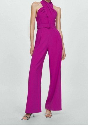 Donna con una tuta smanicata colore magenta con scollo halter, pantaloni a gamba larga e cintura abbinata, abbinata a tacchi aperti argentati.