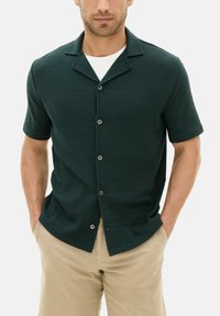 Tummanvihreä lyhythihainen nappipaita, jossa on teksturoitu kangas, kaulukseton kaula-aukko ja viisi mustaa nappia edessä.