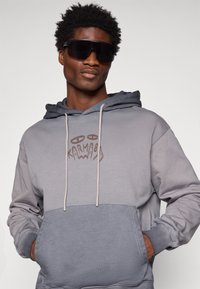 Sivi dukserica s dvobojnim dizajnom, s kapuljačom na šnuru i prednjim džepom. Istaknuti grafički natpis "KARMA" u smeđoj boji na grudima.