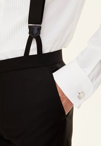 Schwarze Hose mit Hosenträgern, weißes Hemd mit strukturiertem Manschetten und silberner Manschette, Hand in der Hosentasche, polierte Oberfläche.