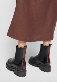 Bottes de cheville en cuir noir avec un motif crocodile texturé, accent rouge brillant à l'arrière et semelle en caoutchouc noire épaisse.