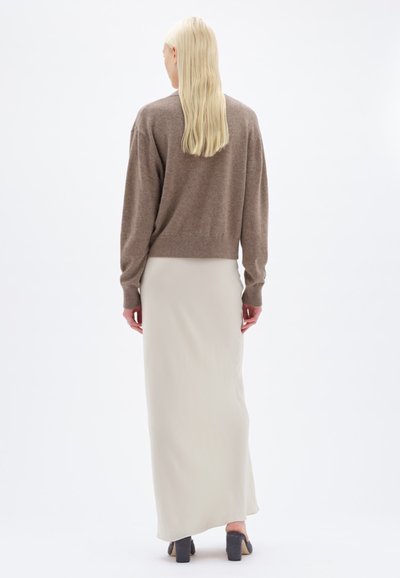 Maglione marrone lavorato a maglia con spalle scese, abbinato a una lunga gonna beige chiara. Il modello indossa scarpe con tacco blocco grigie.