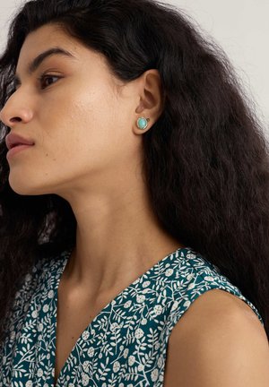 Cercei stud de turcoaz montați în metal cu ton auriu, având o suprafață netedă și formă rotundă. Modelul poartă o rochie verde cu model floral.