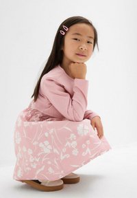 Junges Mädchen mit langen, dunklen Haaren und pinken Haarklammern, das in einem rosa Blumenkleid und beigen Schuhen kauert und die Hand unter das Kinn legt.