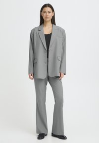 Grå skræddersyet jakkesæt med en lang blazer med hakkede revers, to forlommer og let udvidede bukser. Sort top under, polerede sko.