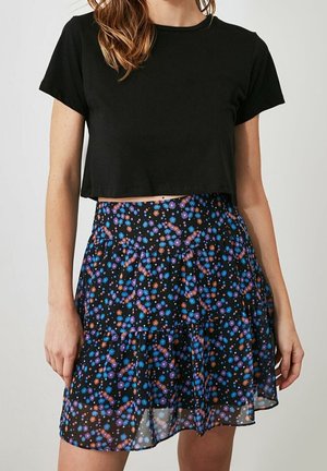 Frau trägt ein schwarzes, kurzes T-Shirt kombiniert mit einem schwarzen Rock, der mit kleinen blauen und orangefarbenen Blumen gemustert ist.