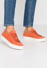 Oranžové semišové tenisky s bílou gumovou podrážkou, vybavené šňůrkami ve stejném odstínu a hladkou texturou. Minimalistický design s nízkým profilem.