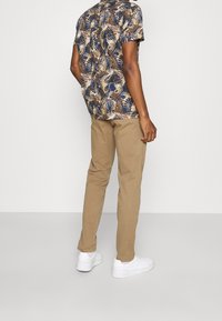 Man med kortärmad skjorta med tropiska lövtryck, beige byxor och vita sneakers som står mot en enkel ljus bakgrund.