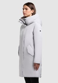 Šviesiai pilkas ilgas paltas su aukštu apykakliu, spaudžiamais užtrauktukais ir dviem šoninėmis kišenėmis. Pagamintas iš lygaus, vandeniui atsparaus audinio.