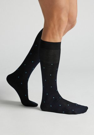 Jambes portant des chaussettes noires montantes avec de petits motifs d'ancres bleues, debout sur un sol gris clair contre un fond uni.