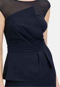 Une robe bleu marine qui présente un corsage ajusté en tissu transparent, un panneau asymétrique, une taille peplum et un détail ceinturé sur le côté.