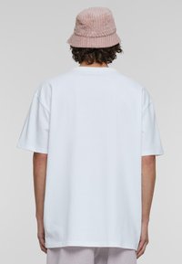 Balta per didelė t-shirt su trumpomis rankovėmis, turinti apvalią iškirptę. Dėvima su rožine banguota skrybėle, demonstruojančia tekstūrą ir švelnumą.