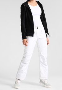 Zwarte zip-up jas, witte aansluitende shirt, witte sneeuwbroek en grijze veterschoenen. Soepele materialen, functioneel ontwerp en contrasterende kleuren.