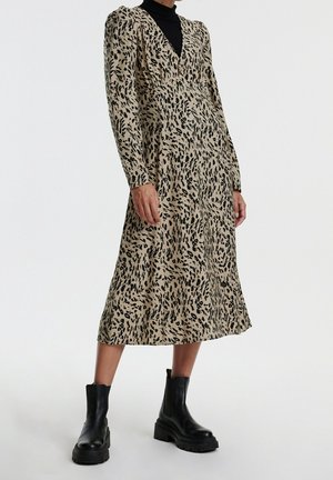 Kvinde iført beige midi-kjole med sort uregelmæssigt leopardmønster og sorte ankelstøvler, stående mod hvid baggrund.