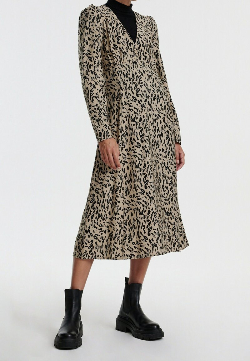 Femme portant une robe midi beige avec un imprimé léopard irrégulier noir et des bottines noires, debout devant un fond blanc.