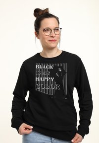 Donna che indossa occhiali e un felpa nera con il testo "Il nero è il mio colore felice" e un'immagine di una persona che tiene un ombrello.