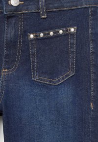 Sötétkék farmernadrág zsebe, a tetején hat ezüst szegecsekkel és kontrasztos narancssárga cérnával körbevarrva.