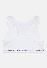 Sostén deportivo blanco con diseño de espalda nadadora y banda elástica, marcado con "TOMMY HILFIGER" en letras rojas y azules. Textura de tela suave.