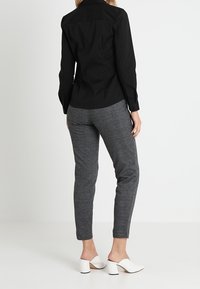 Chemise noire ajustée à manches longues, pantalon gris fuselé, et mules blanches à talons, vues de dos sur un fond blanc uni.