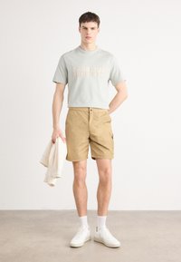 Tricou verde deschis cu logo-ul "BARBOUR INTERNATIONAL", pantaloni scurți bej, teniși albi, ținând un hanorac de culoare deschisă.