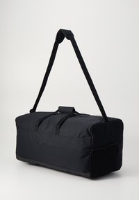 Μαύρη τσάντα duffel από ανθεκτικό ύφασμα, με φερμουάρ στο πλάι, δύο χερούλια και ρυθμιζόμενο λουρί ώμου.