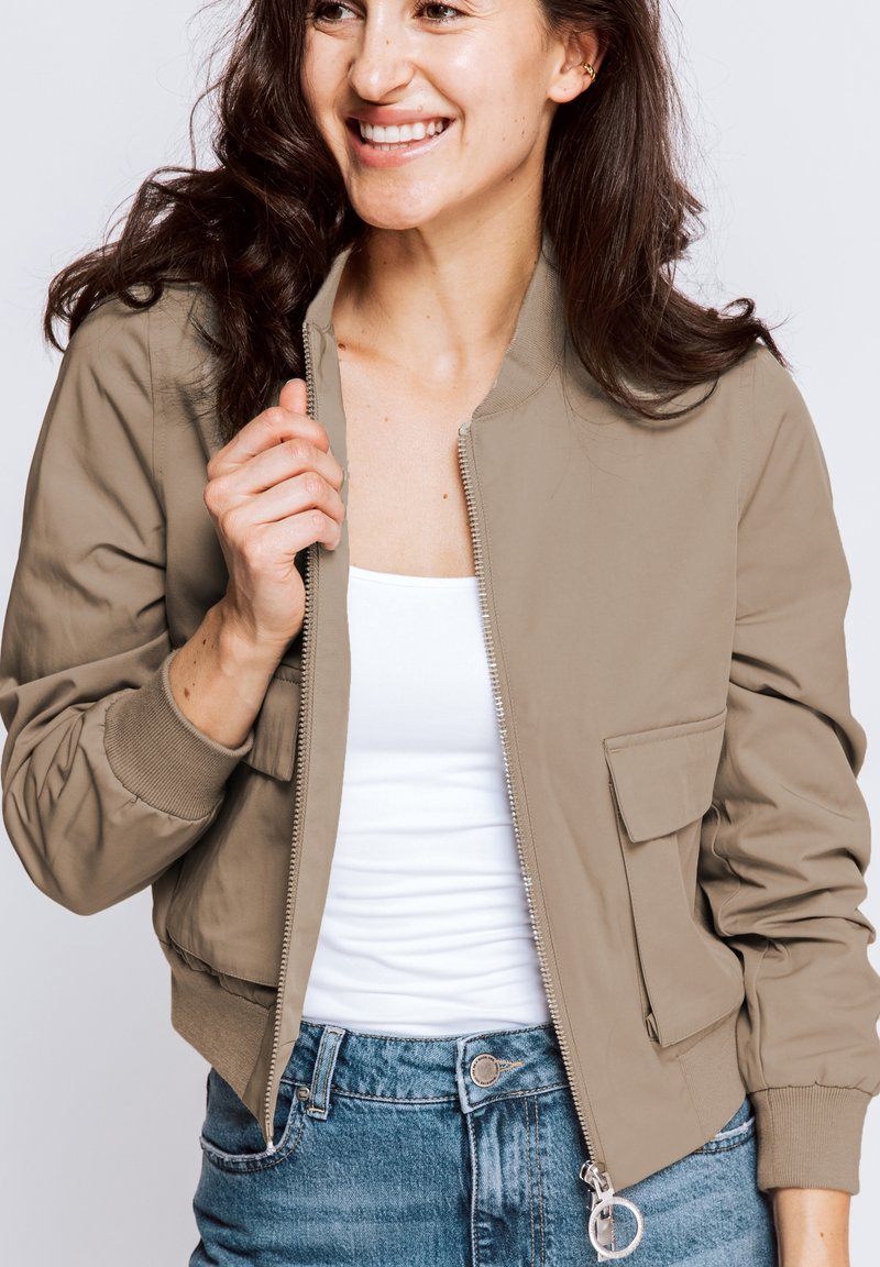 Beige bomber jacket with a zip front, ribbed cuffs, and a pocket on the left. Worn over a white top, paired with denim jeans.