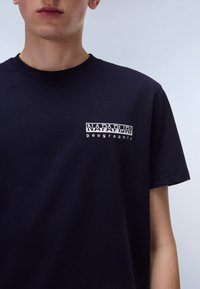 Camiseta de algodón azul marino con un logo en el pecho que dice "NAPAPIJRI" y "geographic" en texto blanco. Cuello redondo sencillo y mangas cortas.