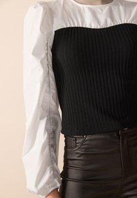 Haut noir à côtes avec des manches ballons blanches, présentant un col incurvé et un tissu texturé, associé à un pantalon noir ajusté brillant.