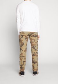 Camisa blanca de manga larga combinada con pantalones cargo de camuflaje que cuentan con múltiples bolsillos y un corte relajado, llevados con zapatillas negras.