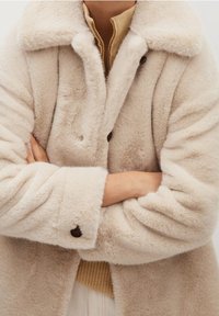 Kræmfarvet falsk pelsfrakke med krave, store knapper og rullede ærmer, lagdelt over en beige strikbluse. Blød tekstur synlig.